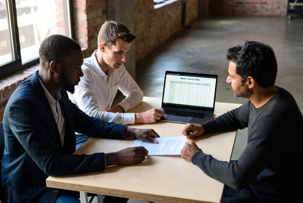 Three founders around a table reviewing a printed term sheet and an ownership table on a laptop