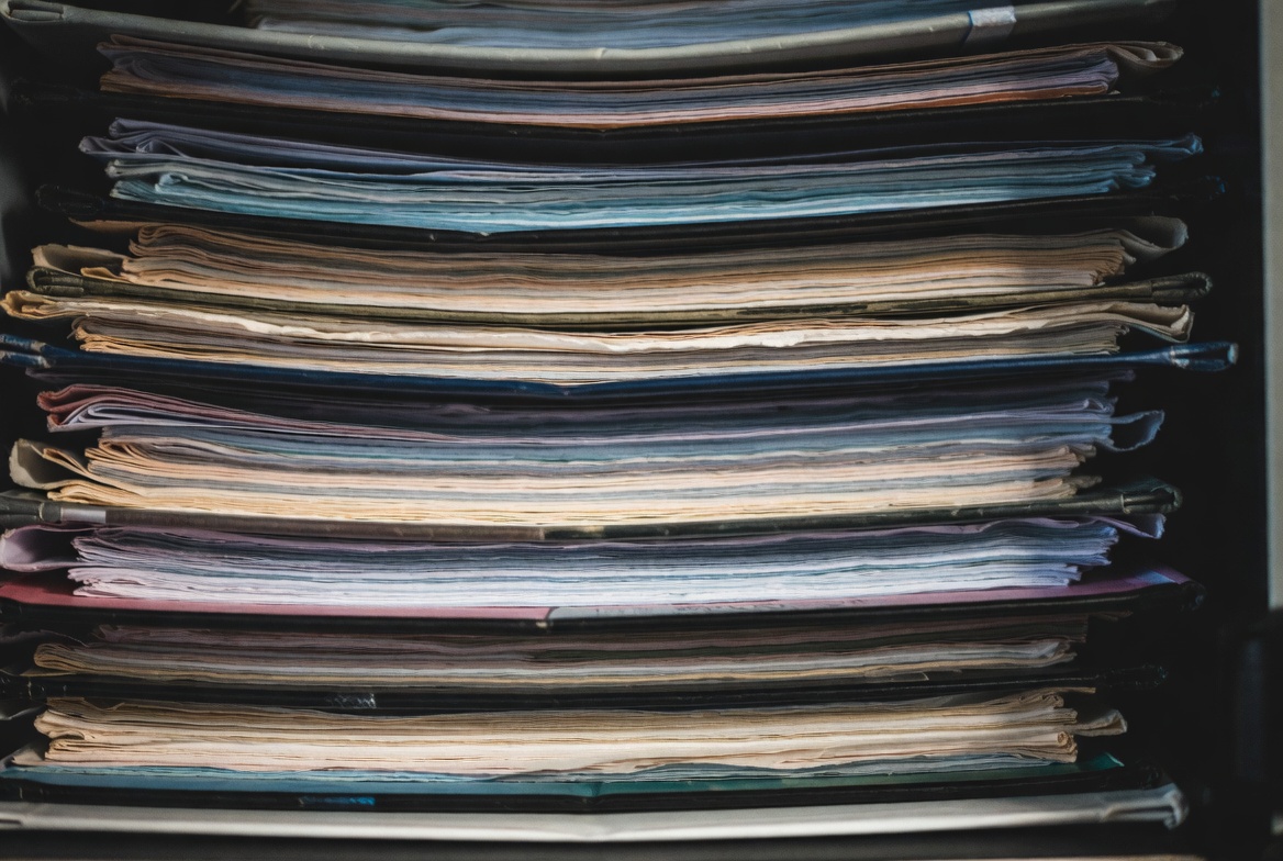 A dense stack of used binders, folders and paper seen close up, edges worn from repeated handling